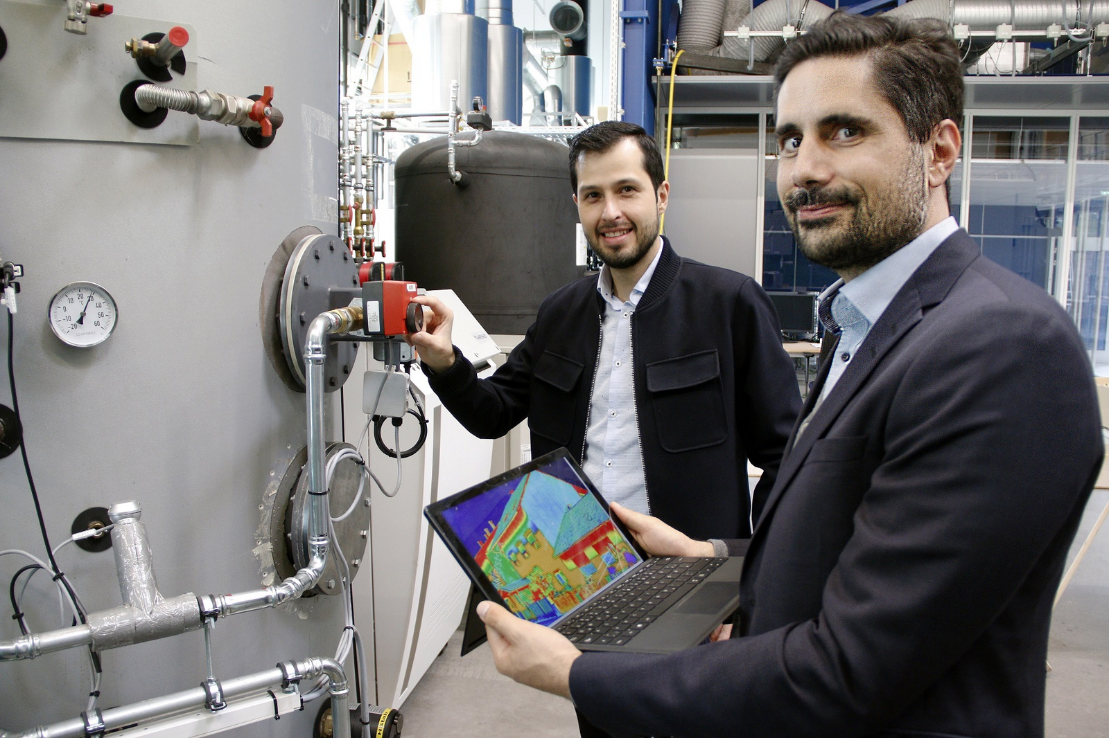 Prof. Dr. Rainer Gasper (rechts) und Mitarbeiter Oscar Villegas Mier (von rechts) vor einer Heizungsanlage. Prof. Gasper hält ein Laptop in den Händen auf dem die wärmeverteilung in einem Haus zu sehen ist.